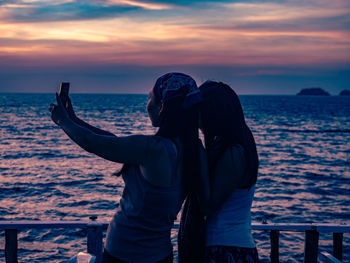 Rear view of 2 women taking a selfie against sky during sunset
