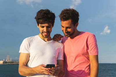 Happy hispanic sportsman in pink t shirt leaning on shoulder of friend using cellphone against blue sky and sea during break in fitness workout