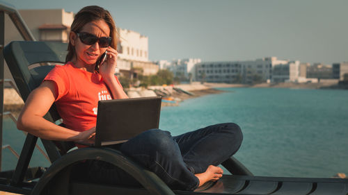 Woman talking on mobile phone while working on laptop against lake