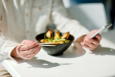 Midsection of person holding chopsticks and phone by food in restaurant