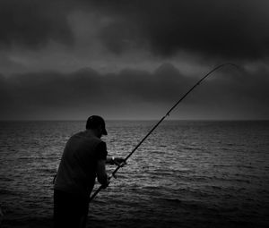 Man fishing in sea against cloudy sky