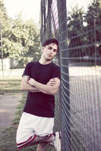 Portrait of man with arms crossed while leaning on chainlink fence at field