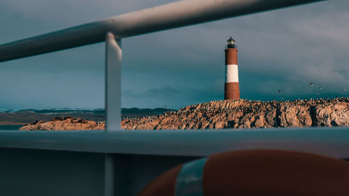 Lighthouse on rock by sea against sky seen through boat railing