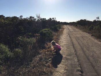 Side view of girl crouching on dirt road by field against clear sky