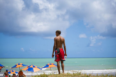 Rear view of shirtless man standing at beach against sky