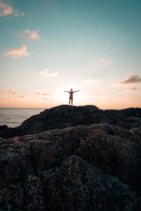 Distant view of person standing on rocky shore against cloudy sky during sunset