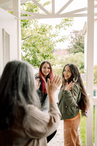 Senior woman waving hand at daughter and granddaughter from entrance of house