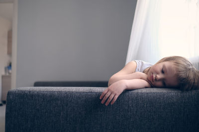 European toddler child sits and gets bored on gray sofa in bright living room, minimalist interior