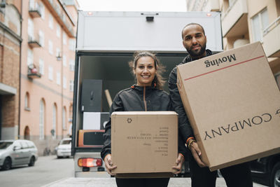 Portrait of male and female messengers carrying cardboard boxes while standing against delivery van in city