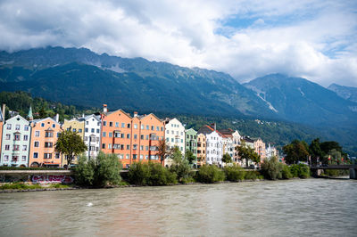 Buildings by river and mountains against sky