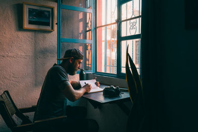 Male sitting at desk writing in a journal illuminated by natural light.
