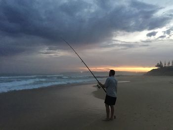 Man fishing at beach against sky during sunset