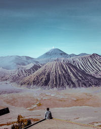 Rear view of man looking at volcanic crater while sitting against sky during sunny day