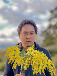 Portrait of young man holding golden wattle flowers against cloudy blue sky and trees.