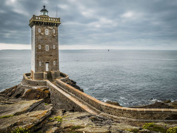 Stone lighthouse by sea against cloudy sky