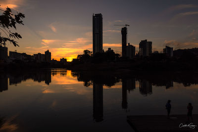 Silhouette buildings by lake against sky during sunset