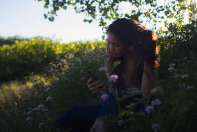 Woman using phone while sitting on field by flower