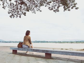 Woman looking at lake while sitting on bench against sky