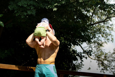 Shirtless father playing with son while standing against branches