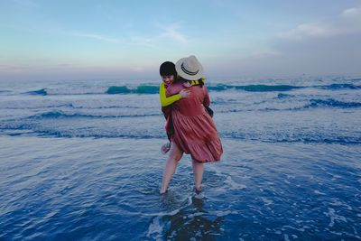 Mother carrying son while standing at beach against sky