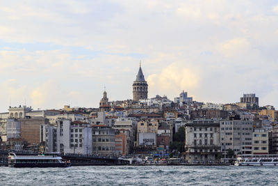 Galata tower amidst buildings in front of sea against cloudy sky