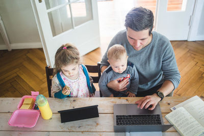 High angle view of father working on laptop while sitting with cute son by daughter using digital tablet at table