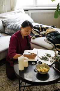 Young woman writing in book while sitting with candles and herbs at table
