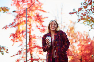 Low angle portrait of smiling young woman holding disposable cup while standing against trees at park during autumn