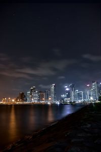 Illuminated buildings by river against sky at night