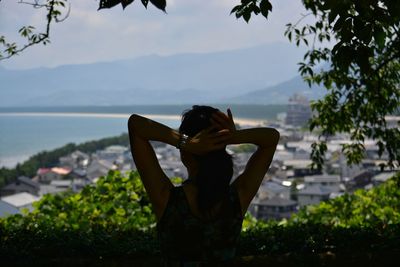 Back view of woman with arms raised standing by sea against sky overlooking city