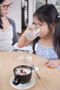 High angle view of girl drinking water while sitting by coffee cup on table with mother in background