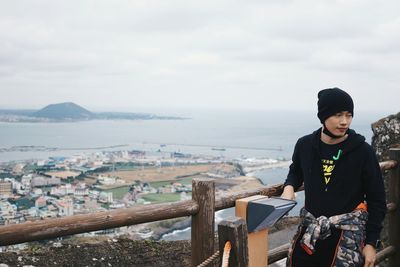 Young man in warm clothing standing by railing against sea