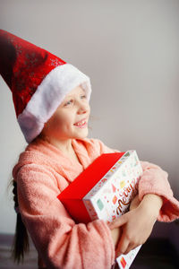 A caucasian girl in a red santa cap happily holds a gift box on a light background, quarantine.