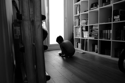 Shirtless boy crouching on hardwood floor by shelves at home