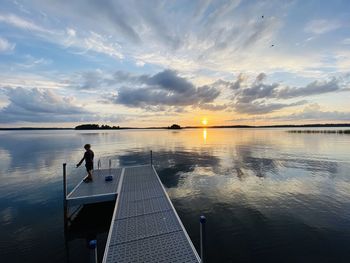 Boy standing by lake on pier against sky during sunset