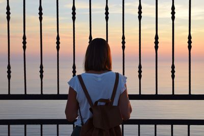 Rear view of woman looking at sea through railing against sky during sunset