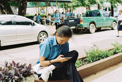 Young woman using mobile phone while sitting on footpath in city