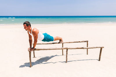 Man exercising on parallel bars at beach against blue sky