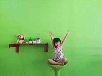 Smiling girl with arms raised sitting against green wall