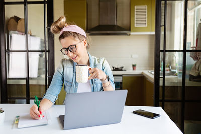 Mid adult woman writing in diary while holding coffee cup at home