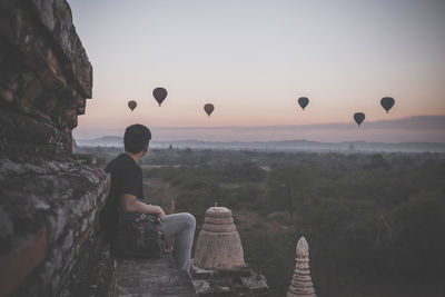 Side view of man looking at hot air balloons flying over landscape during sunset