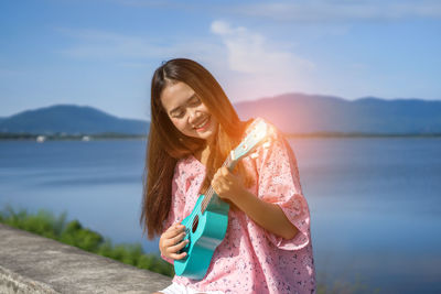 Smiling young woman playing ukulele against lake and sky