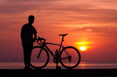 Silhouette man standing by bicycle against orange sky