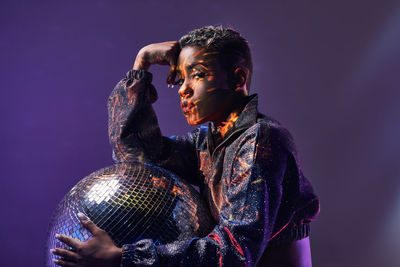 Side view of trendy young african american female with makeup looking at camera while holding glitter ball in violet light