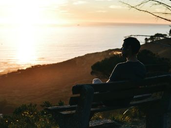 Rear view of man smoking while sitting on bench against sea during sunset