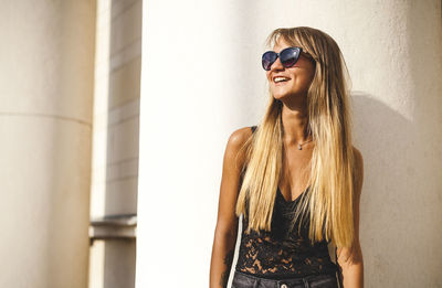 Portrait of smiling young woman wearing sunglasses standing against wall