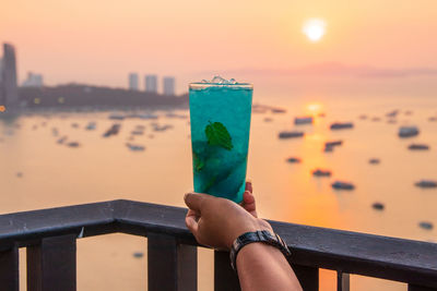 Person holding glass of railing against sea during sunset