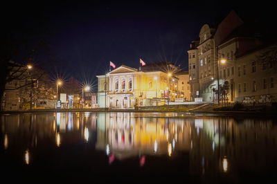 Illuminated buildings by lake against sky in city at night