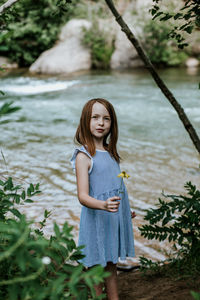 Vertical portrait of a young girl holding a flower next to a river