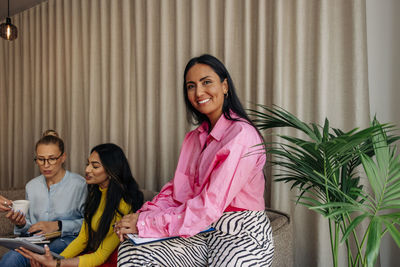 Portrait of smiling businesswoman sitting near plant with colleagues at office